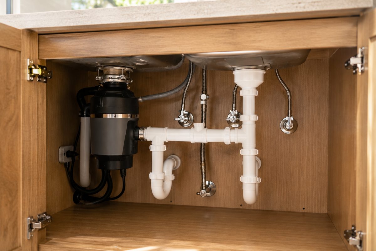 Under-sink view of clean new PVC drain lines, shutoff valves, supply lines, and a garbage disposal